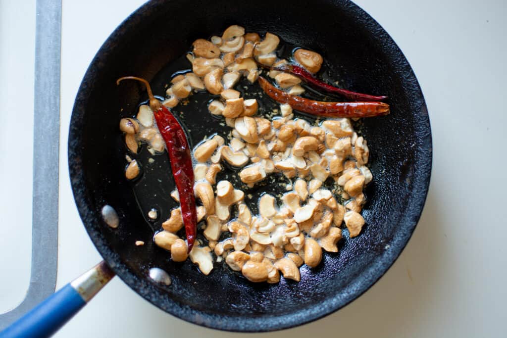 Chopped cashews in a small black frying pan with whole, dried red chiles sitting on a white counter top. 