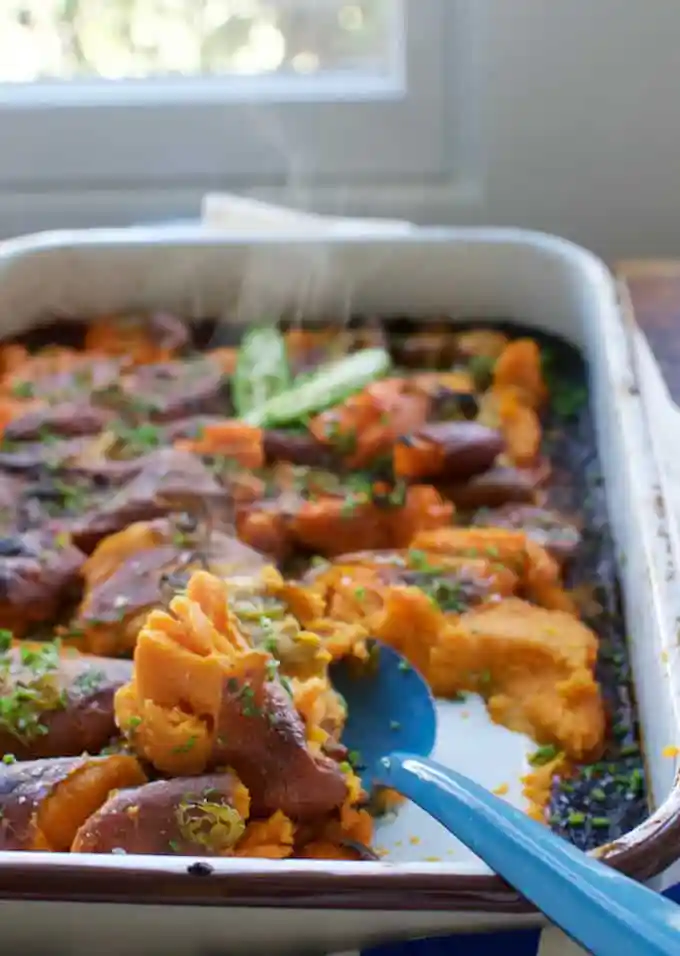 A white casserole dish that has sweet potatoes that are hot and steaming. There is a blue spoon that is taking a scoop out. 