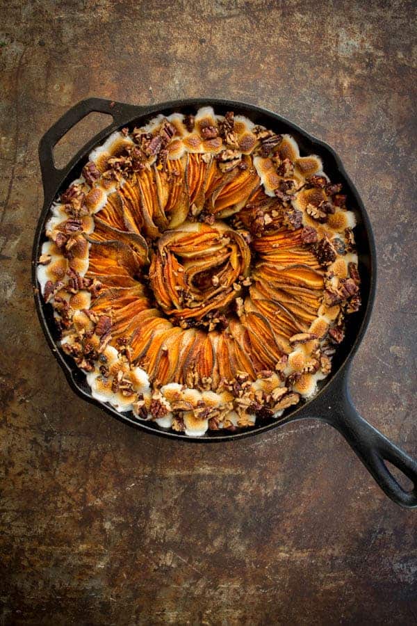 A black cast iron skillet filled with thin slices of sweet potato and apple and topped with marshmallows and pecans. 