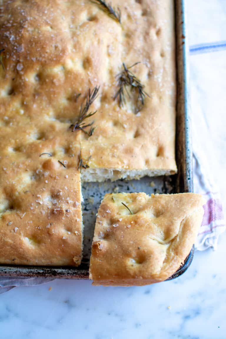 A loaf of focaccia topped with rosemary sitting on a marble table with a piece pulled out from the pan.