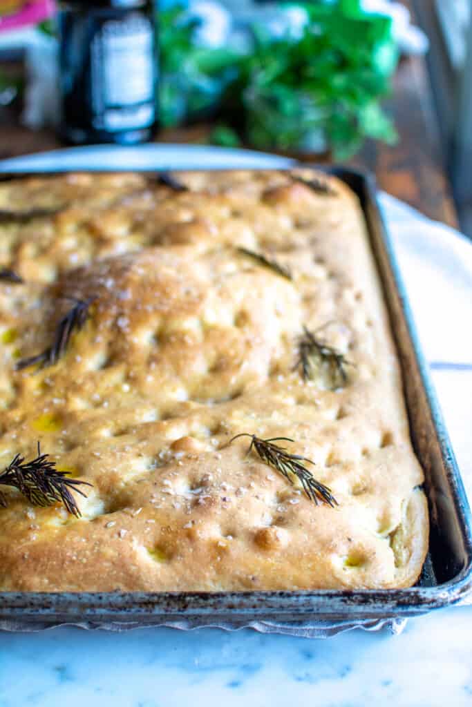 A baking sheet with focaccia in it sitting on a marble table. There is sprigs of rosemary on top and it is sprinkled with salt. 