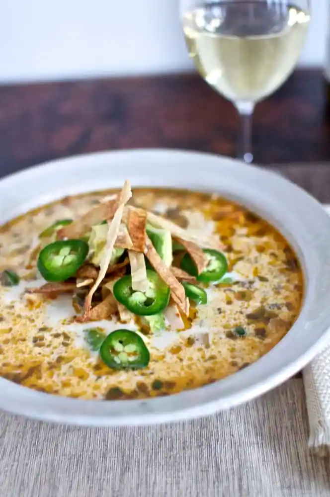 A white bowl filled with creamy chicken tortilla soup topped with tortilla strips and jalapeno slices with a glass of wine. 