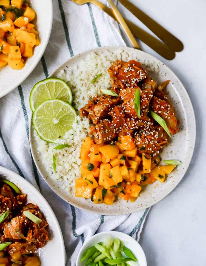 A white plate filled with steamed rice, shredded chicken, mango salsa, and two lime slices sitting on a stripped tablecloth. 