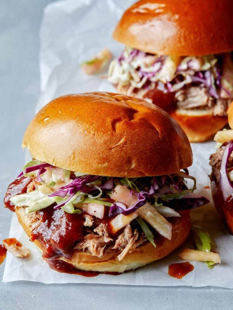 Three pulled pork sandwiches sitting on a piece of parchment paper topped with red cabbage slaw. 