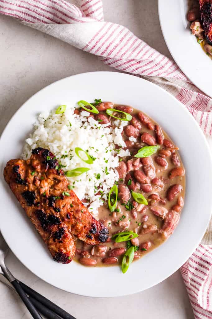 A white bowl of red beans and rice with a side of grilled andouille sausage and topped with thinly sliced scallions. 