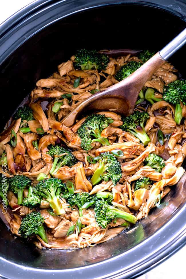 A black crockpot filled with shredded chicken and broccoli with a wood spoon sticking out of one side of the pot. 