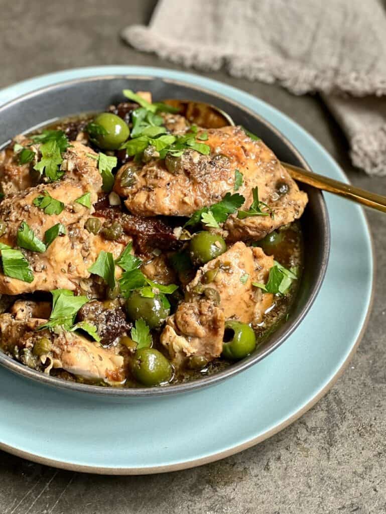 Chicken in a bowl with green olives and parsley leaves on top with a gold spoon in one side of the bowl on a blue plate. 