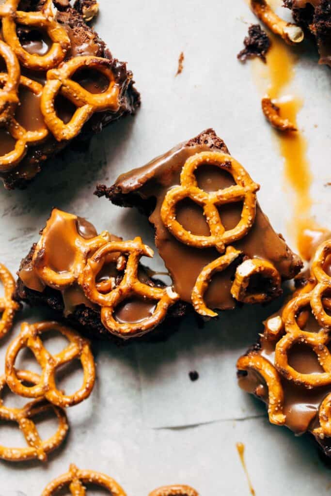 Several brownies sitting on a white background topped with caramel sauce and pretzels, one brownie is broken. 