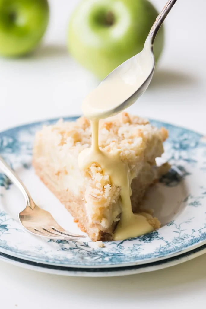 A piece of apple cake on a blue and white plate with a spoon dripping custard sauce on top with green apples behind it. 