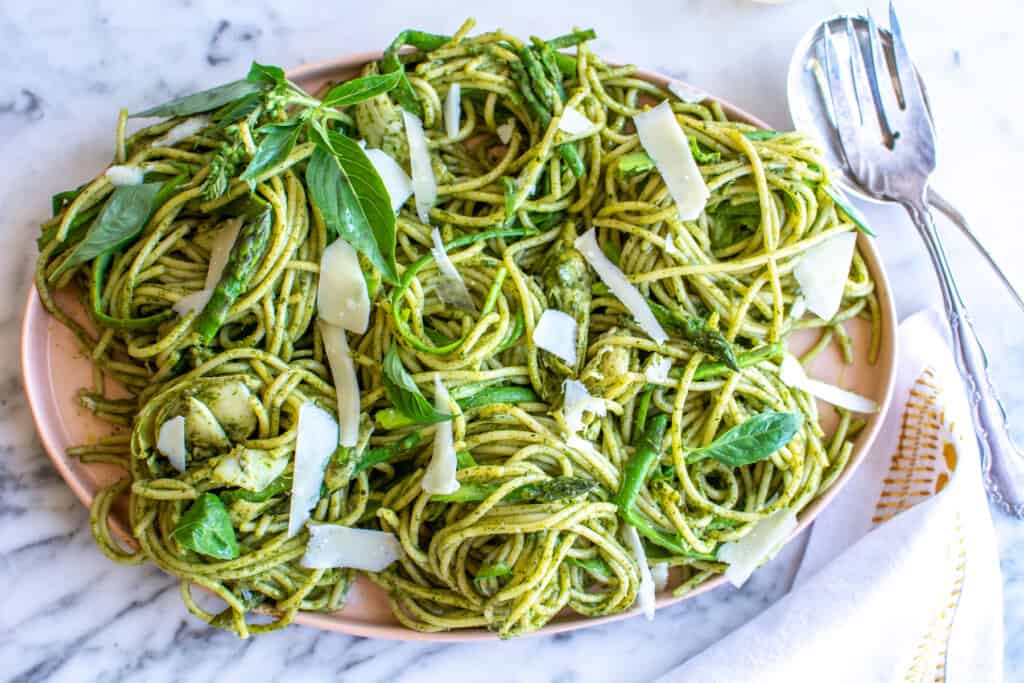 A pink platter of spaghetti with pesto and shaved asparagus on a marble table next to serving spoon and fork. 