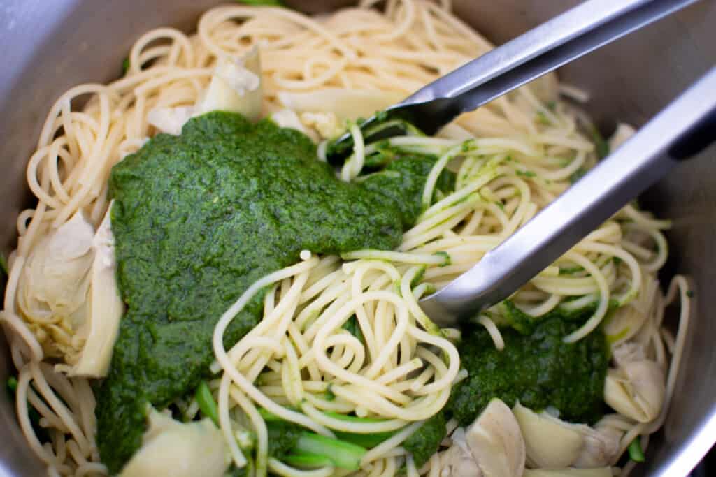 Spaghetti in a pot with green pesto sauce, artichokes and tongs that are stirring the mixture. 