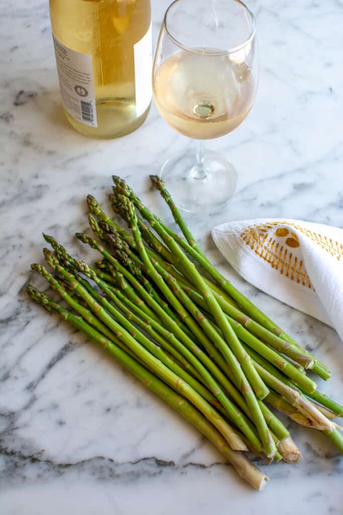 A bottle of white wine with a glass of white wine next to it and a bunch of raw asparagus on a marble table next to a white cloth. 