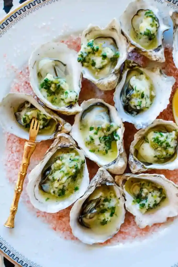 A platter of grilled oysters sitting on a bed of pink salt topped with green Chile garlic butter with a gold fork. 