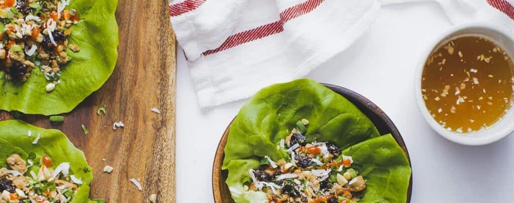 A white table with three plates that have lettuce wraps on them and a small bowl of dipping sauce next to a white towel. 