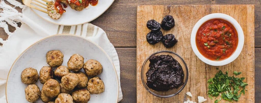 Several turkey meatballs on a plate next to a cutting board of little bowls of prune puree, a white bowl of marinara sauce. 