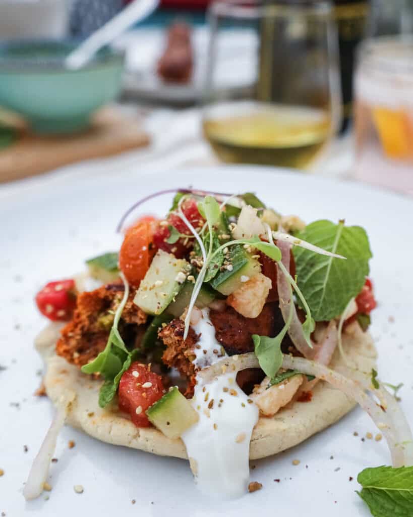 A piece of pita bread topped with ground turkey kofta and salad on top of it on a white plate with a glass of wine behind it. 