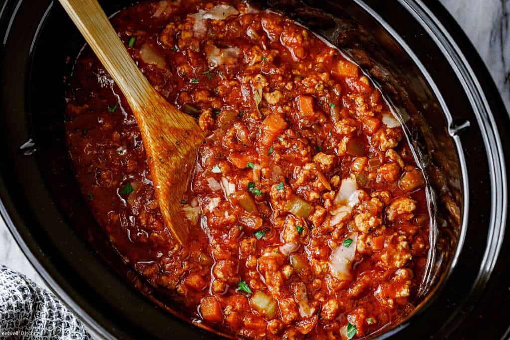 The black container of a slow cooker filled with ground turkey tomato pasta sauce with a wood spoon sticking out of one side. 