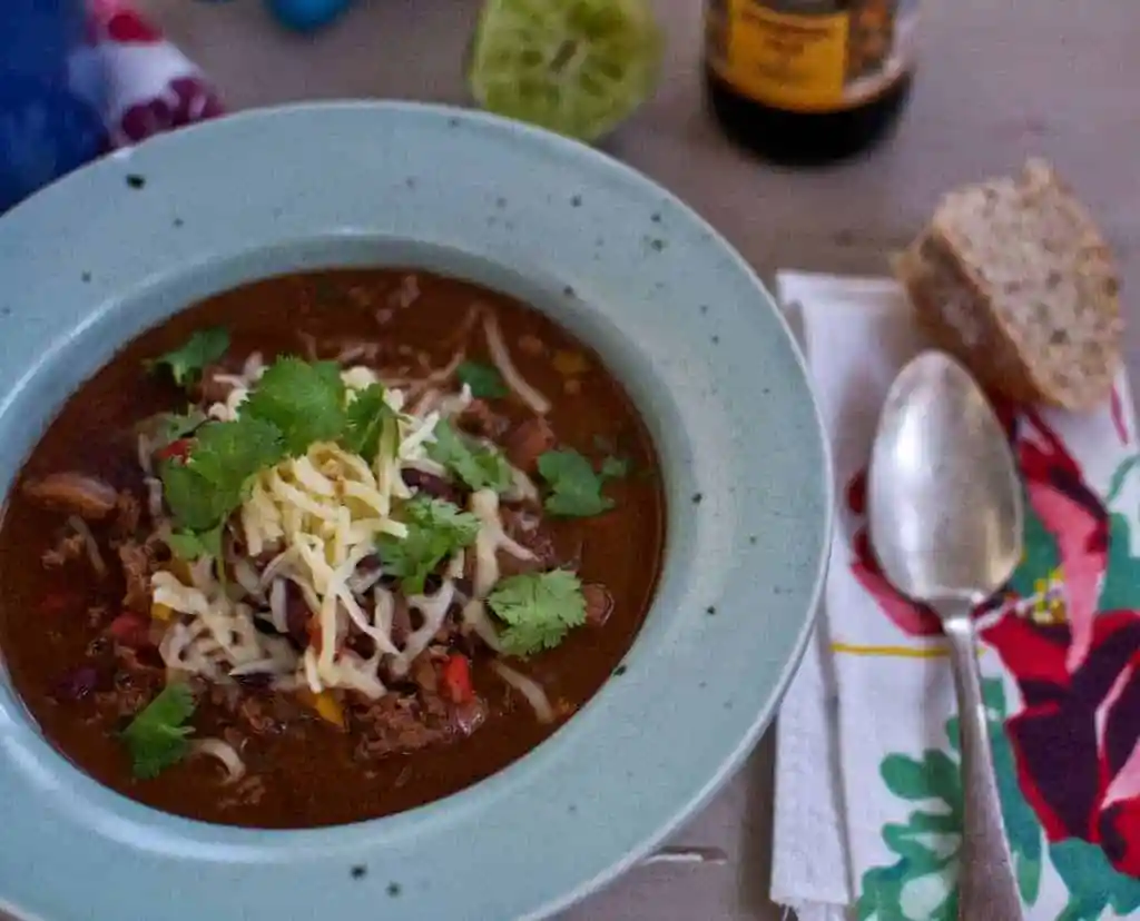 A blue bowl filled with turkey chili with shredded cheese and cilantro leaves on top with a napkin and a spoon. 