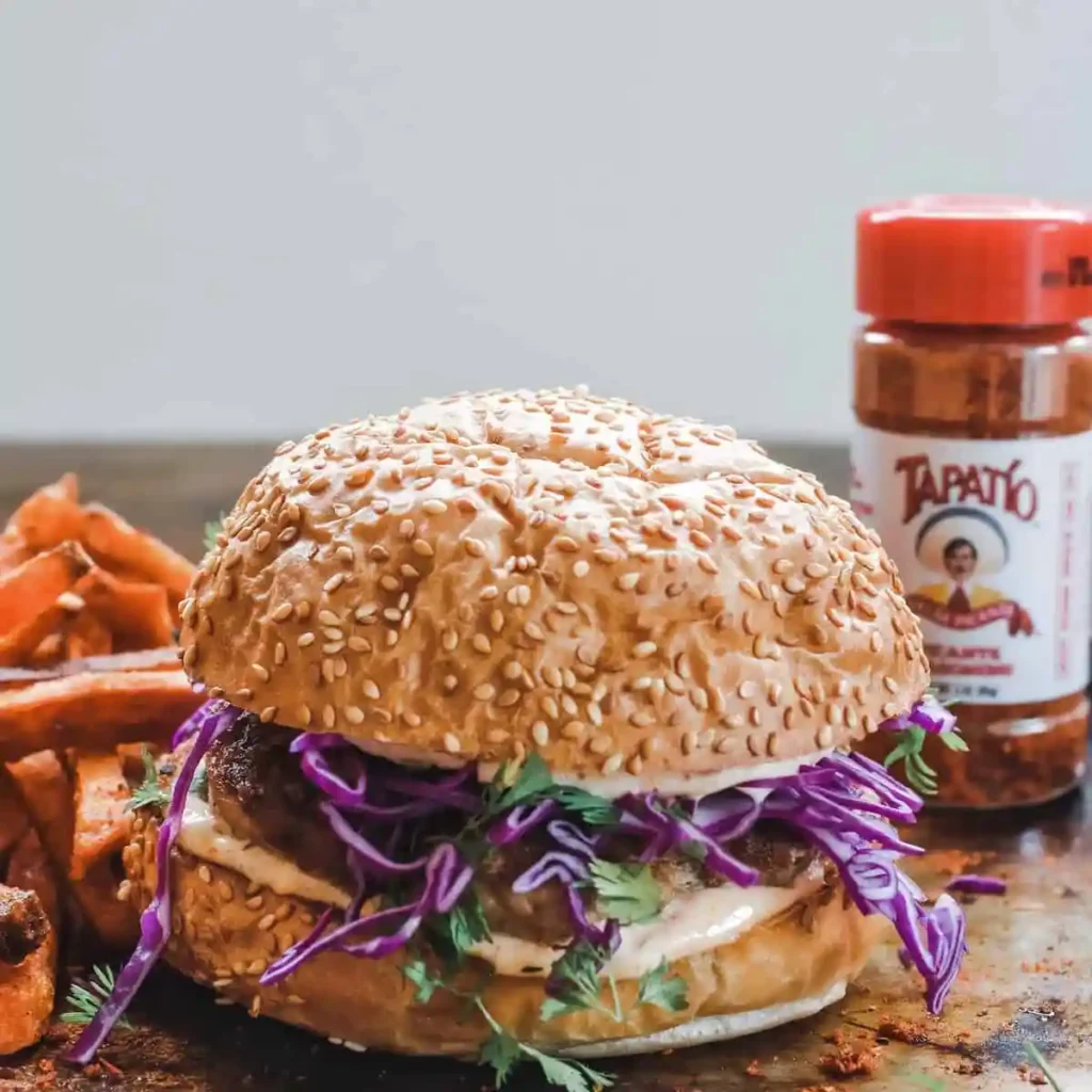 A turkey burger on a wood table with purple slaw and cilantro on top of it with sweet potato fries behind it. 