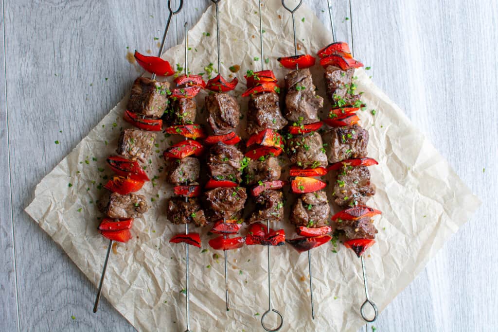 Several beef skewers sitting on a piece of parchment paper on a grey table with chopped bits of parsley on top. 