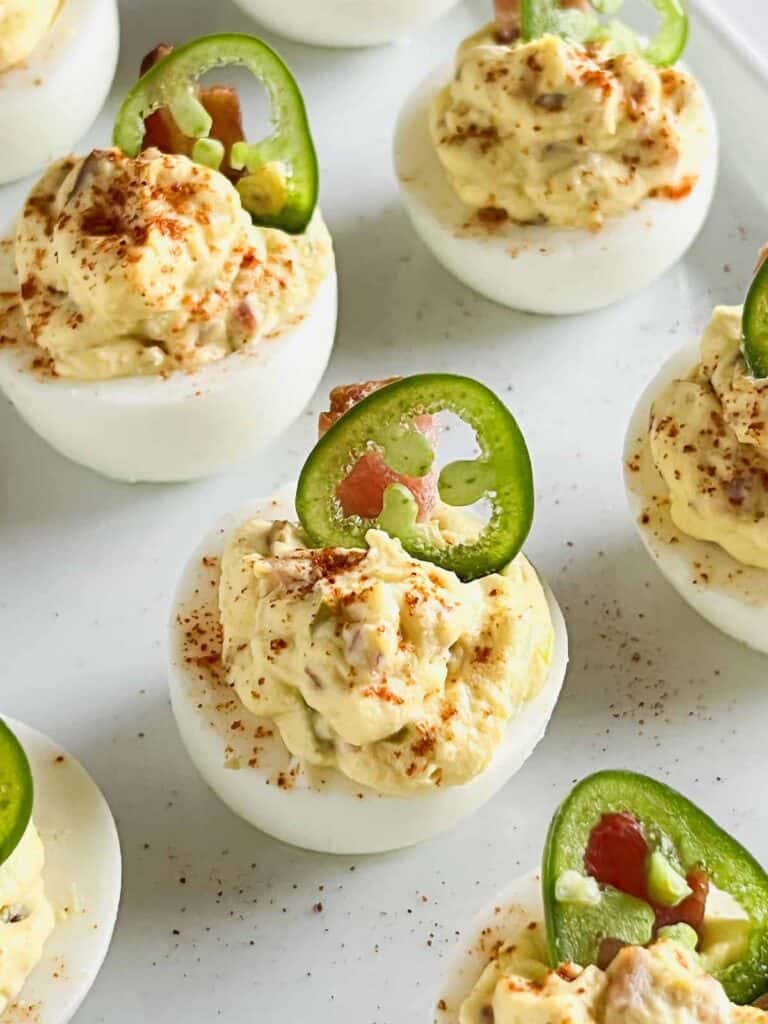 A platter of deviled eggs topped with slices of jalapeño, a piece of bacon and a sprinkle of paprika. 