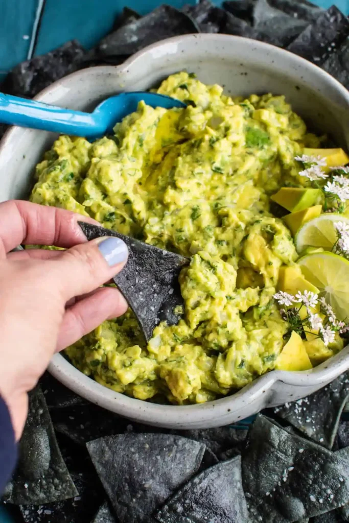 A bowl of guacamole with a blue chip dipping into the bowl and a blue spoon on the side. 