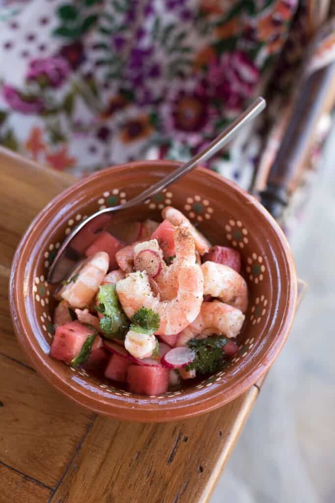 A terracotta bowl of shrimp ceviche with watermelon, cilantro, and sliced radishes with a spoon dipping into one side. 