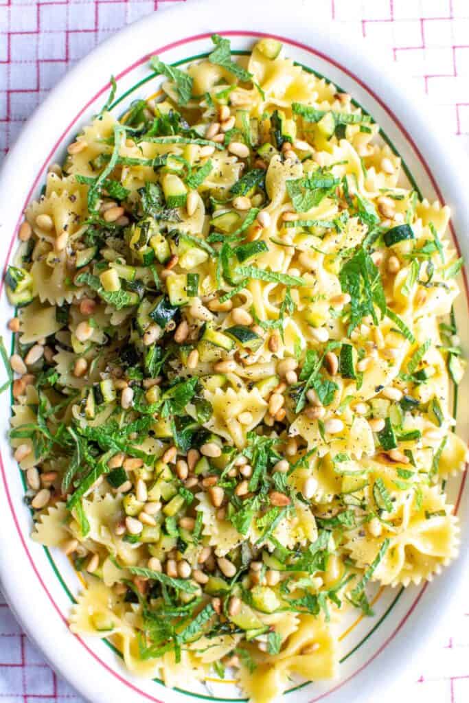 A oval platter filled with bow tie pasta, cubes of zucchini, fresh slices of mint, and toasted pine nuts. 