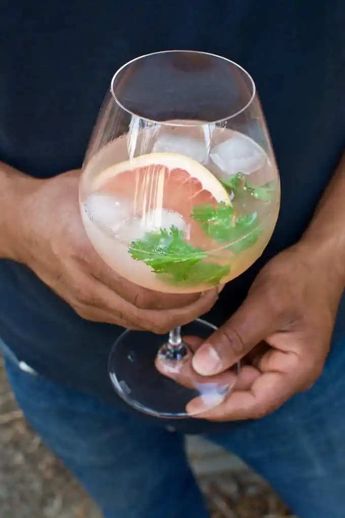 A man holding a wine glass with a cocktail and ice in it and a slice of pink grapefruit and a sprig of cilantro. 