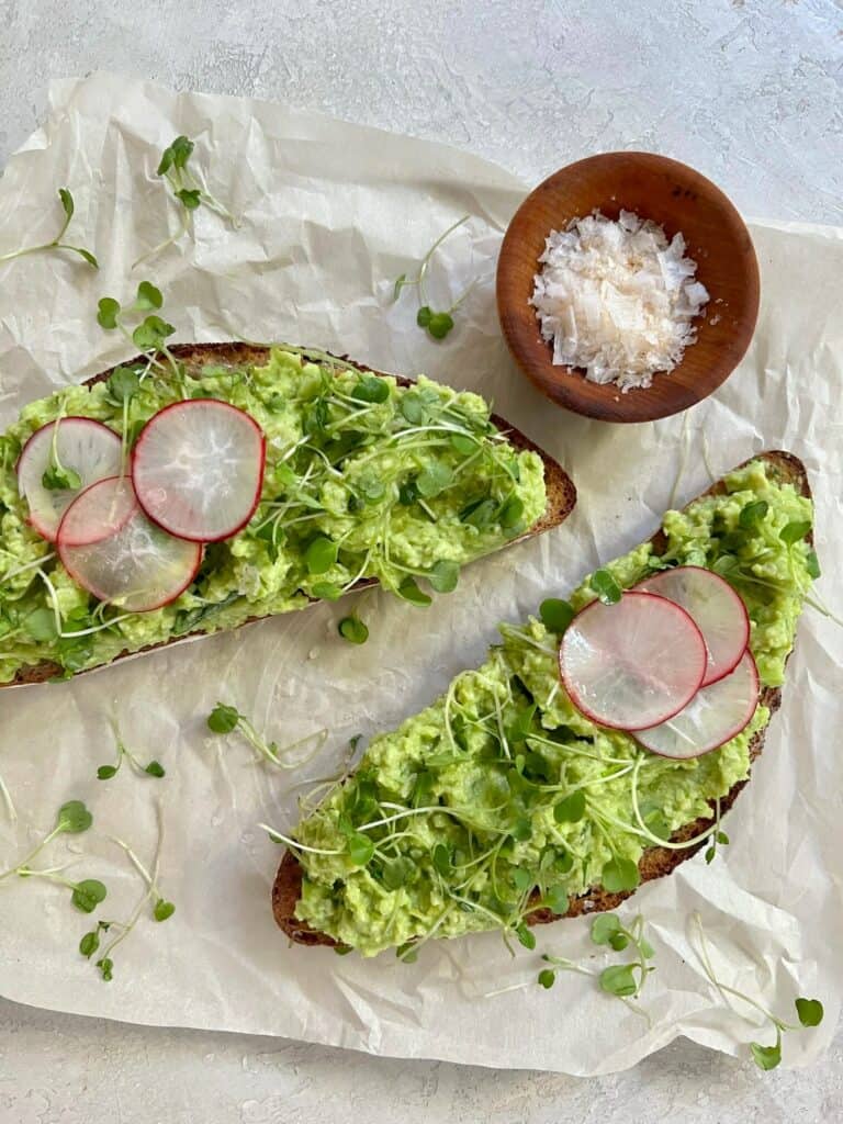 Two pieces of bread spread with avocado and topped with sprouts, radish slices, and sea salt on a piece of parchment. 