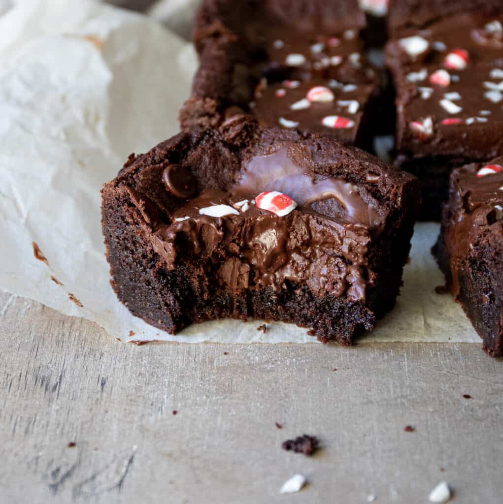 Several brownie squares with a big bite taken out of it and topped with chopped candy canes sitting on parchment paper. 