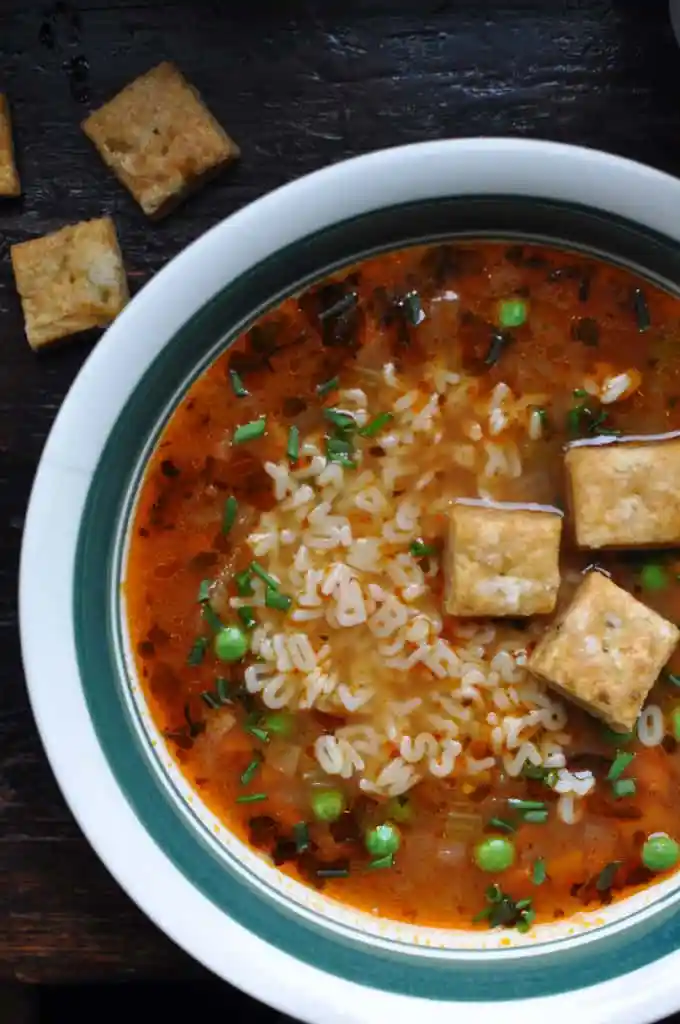 A bowl of vegetable soup filled with alphabet letters and green peas with homemade crackers floating on top. 