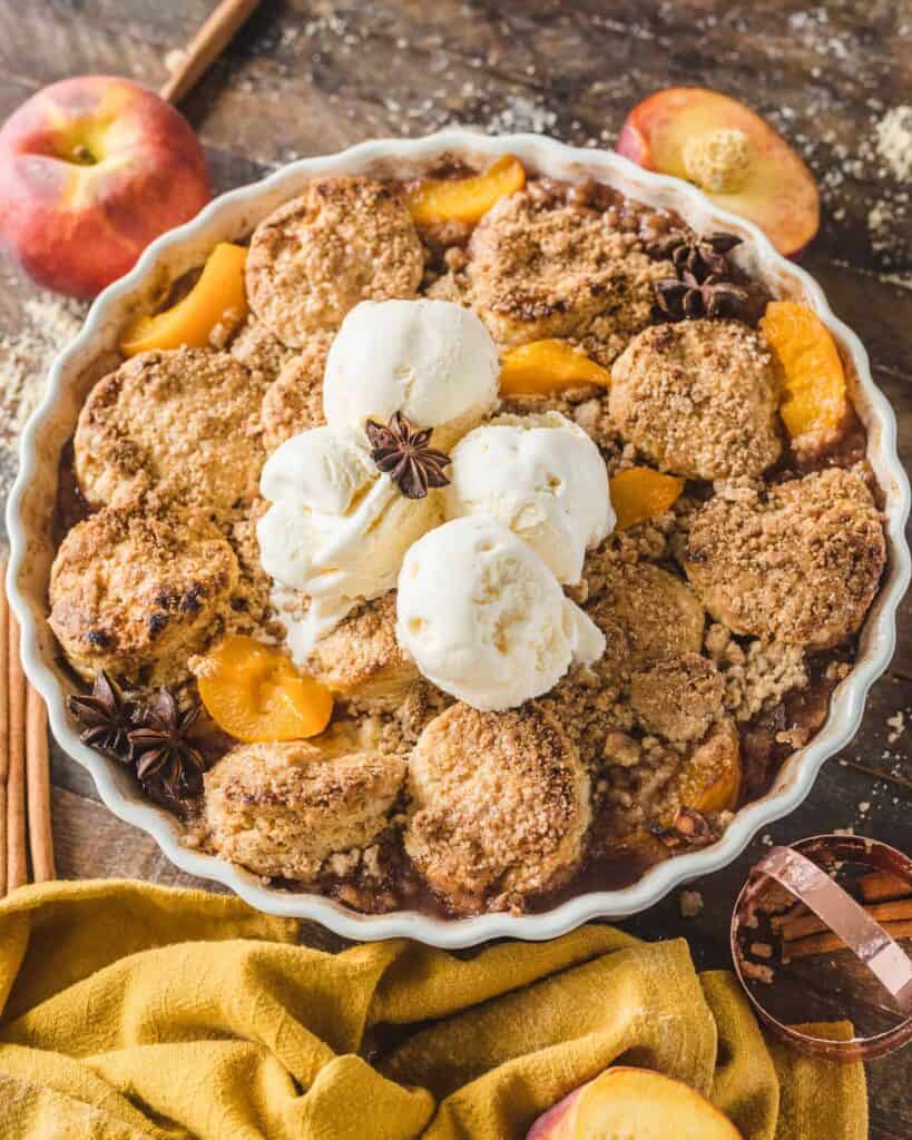 A round dish of peach cobbler topped with flaky biscuits and ice cream and peaches dotting here and there . 