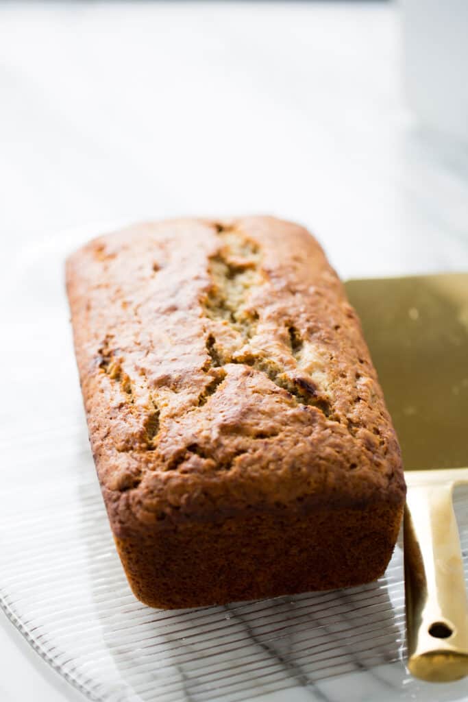 A loaf of banana bread sitting on a glass plate with a bronze knife next to it. 