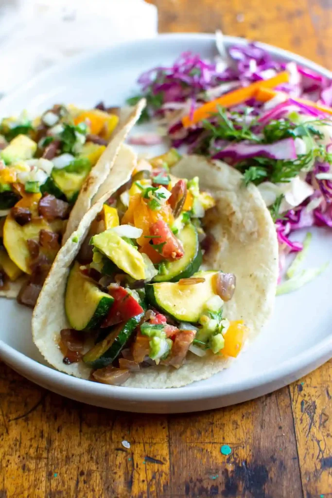 Two squash tacos on a white plate with some slaw on the side on a wood table. 