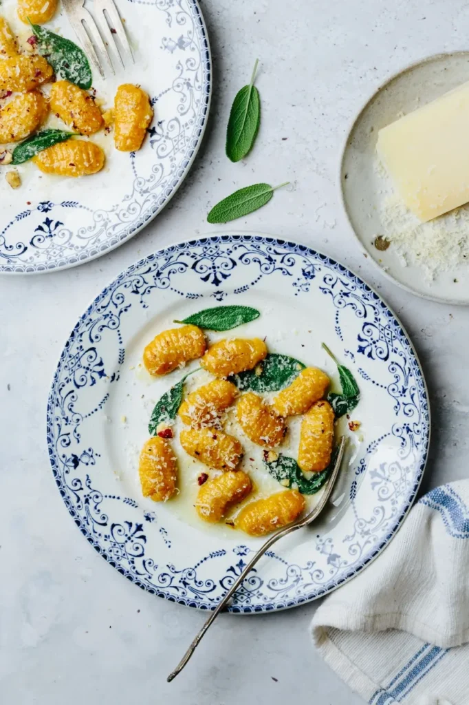 Two plates of gnocchi topped with cheese and sage leaves and a dish of cheese next to them. 