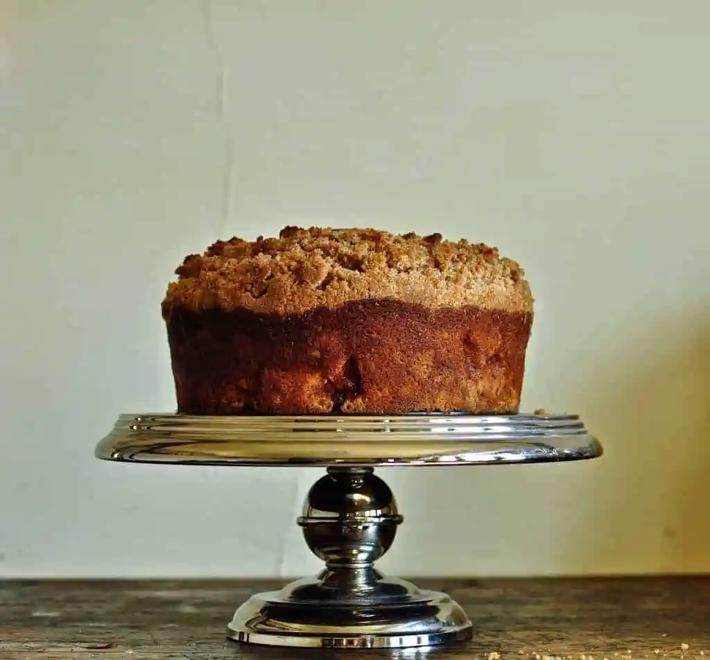 A tall coffee cake on a silver cake stand on a wood table. 