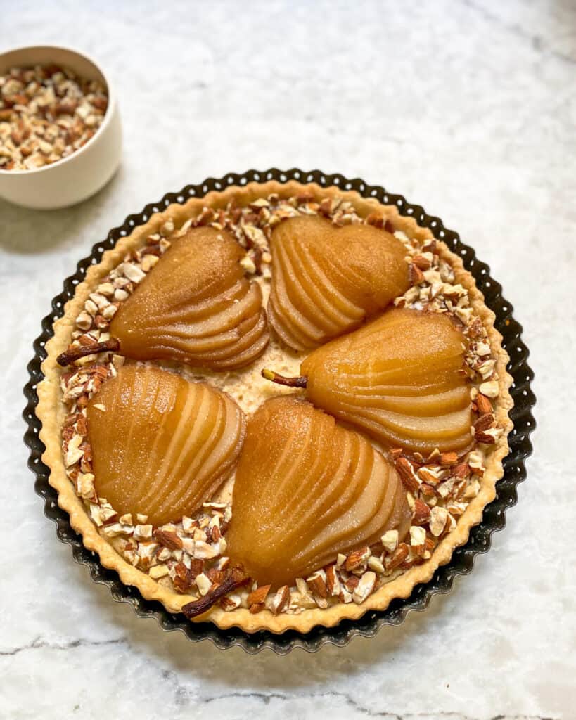 A round tart in a tart tin covered with sliced pears and crushed almonds. 