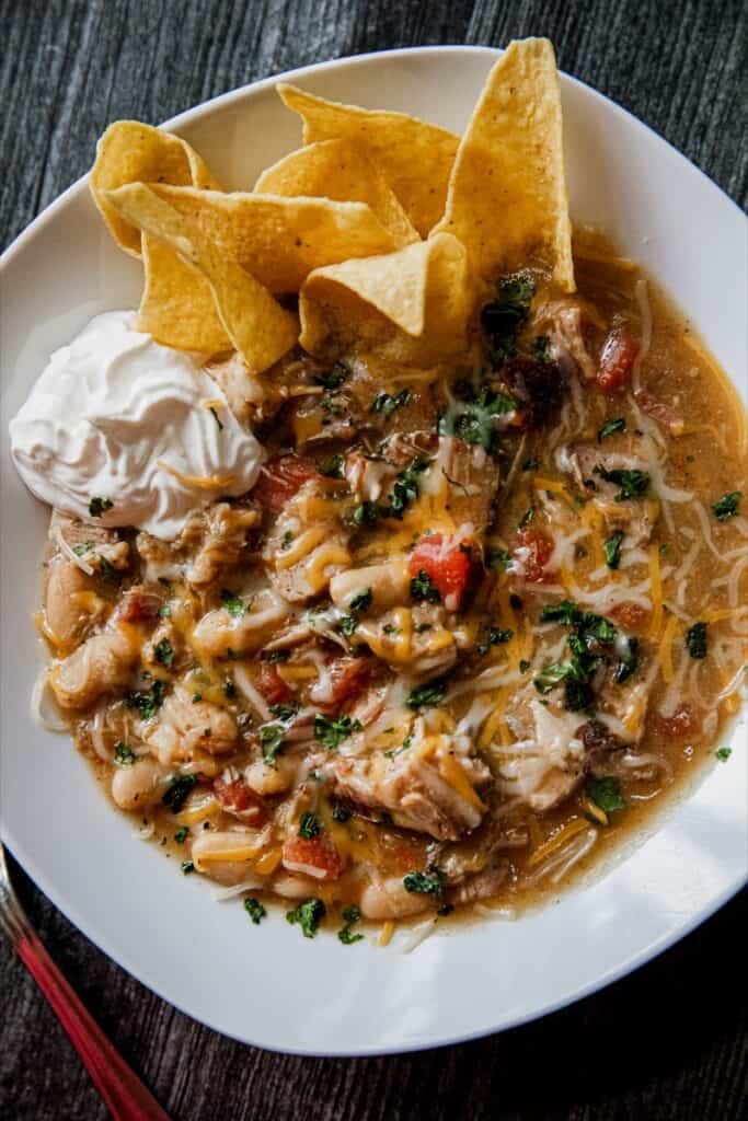 A white bowl with shredded chicken chili topped with chips and a dollop of sour cream. 