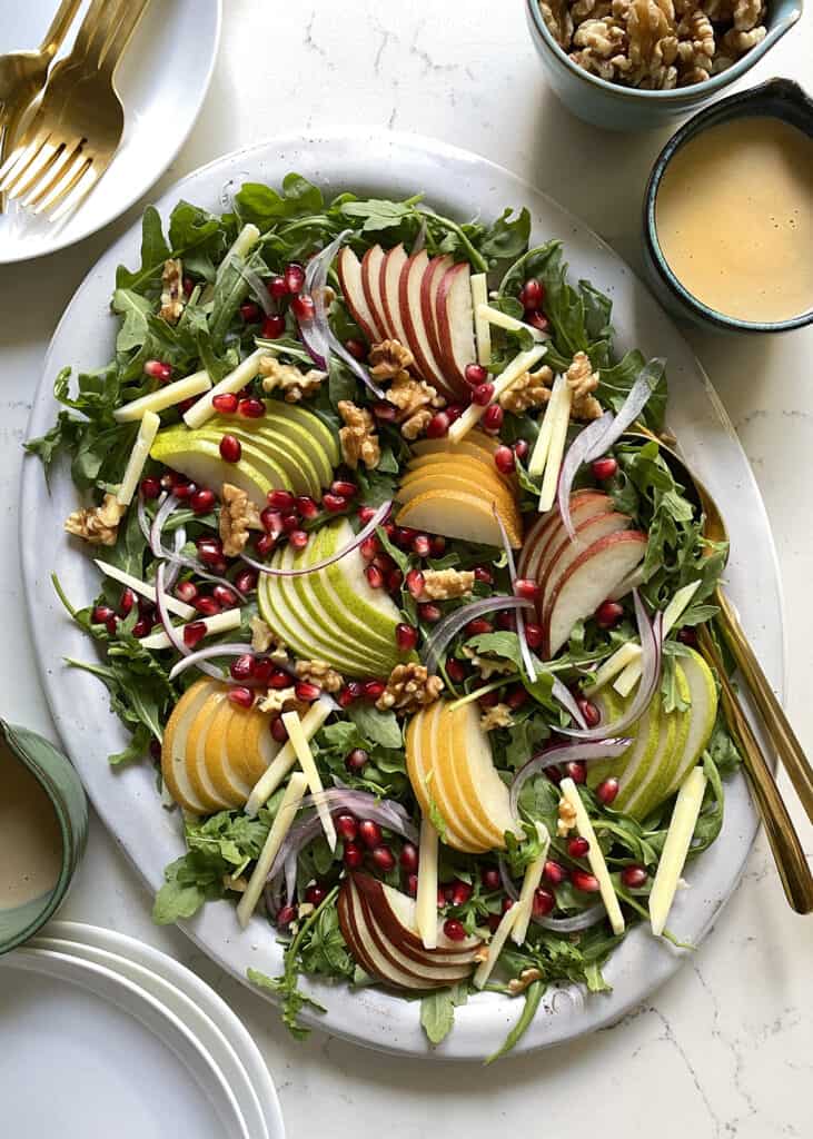 Arugula on a large platter topped with sliced pears, red onions, and walnuts next to a pitcher of dressing and more walnuts. 