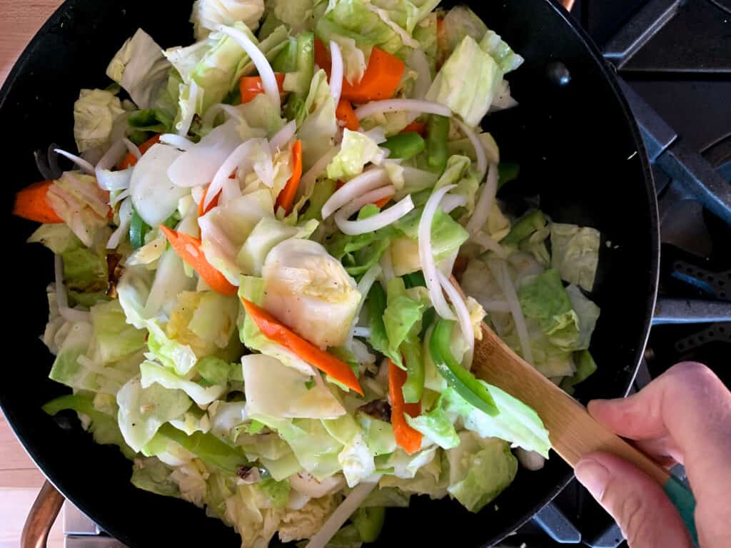 A hand stirring the vegetables with a wood spoon in a black pot. 