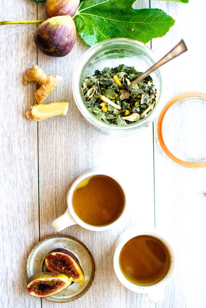 Two cups of fig leaf tea next to a jar of the tea leaves on a table. 