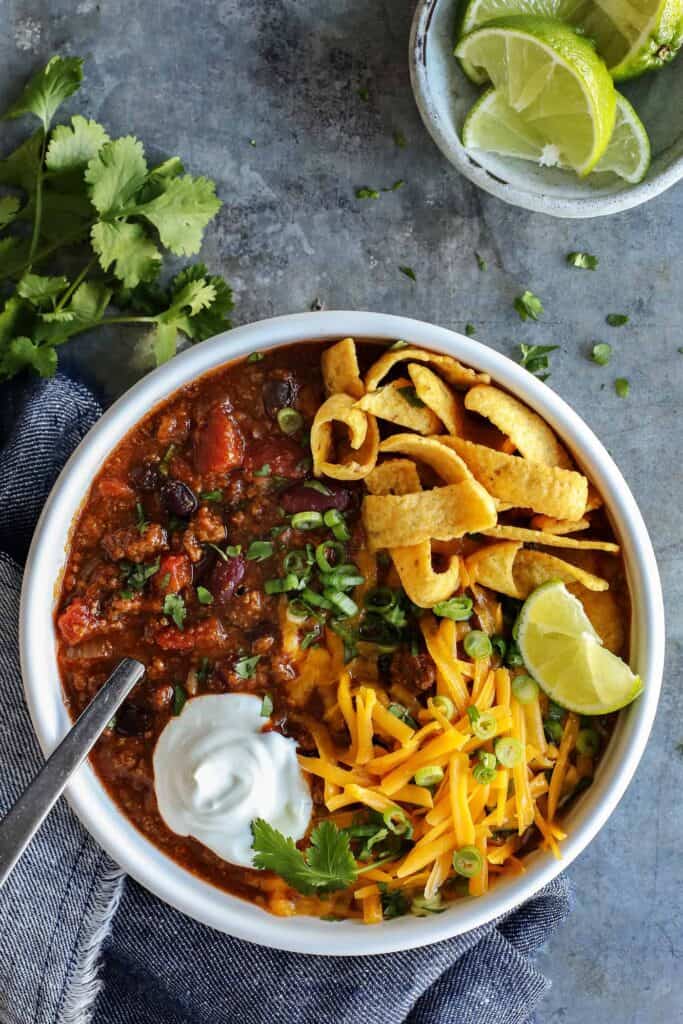 A bowl of chili with tomatoes and red kidney beans topped with Frito chips, shredded orange cheese, and sour cream. 