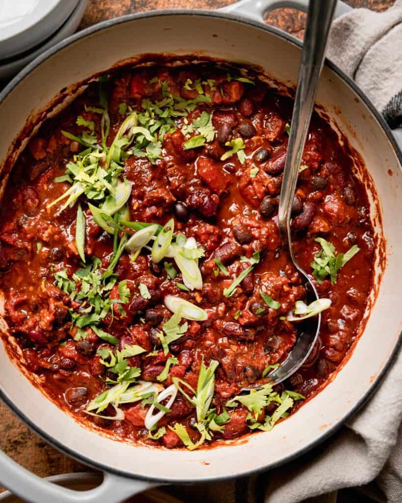 A big pot of red chili topped with thinly sliced scallions and a spoon in the pot. 