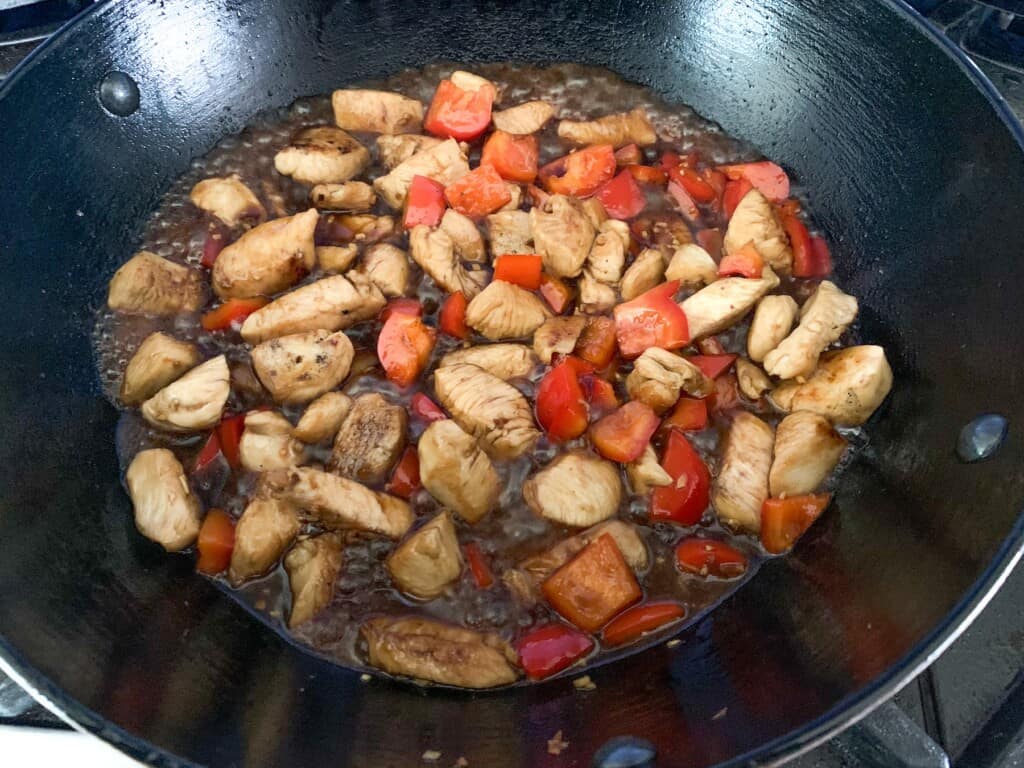 A wok with chicken and red bell peppers and sauce in it. 