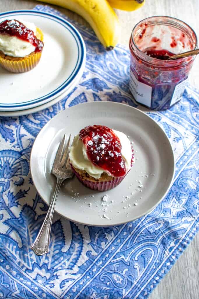 A brown butter banana cupcake on a white plate topped with cream cheese frosting and raspberry jam. 