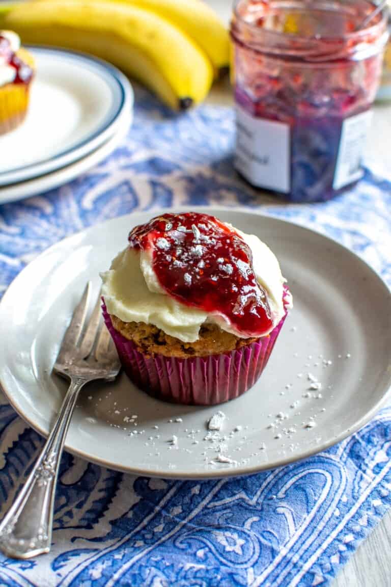 A brown butter banana cupcake on a white plate with a blue tablecloth underneath.