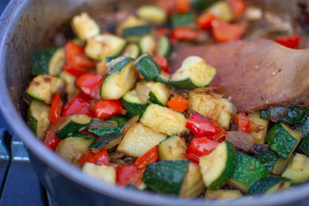 Zucchini and peppers being sautéed in a frying pan with a wood spoon. 