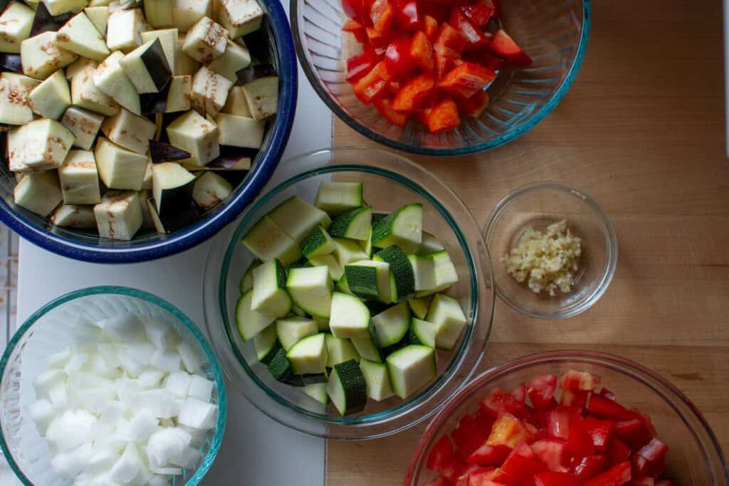 Glass bowls of chopped eggplant, zucchini, tomatoes, and more. 