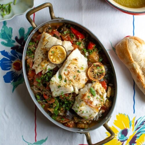 A dish of ratatouille with cod on top and lemon slices next to a small saucepan of butter sauce.